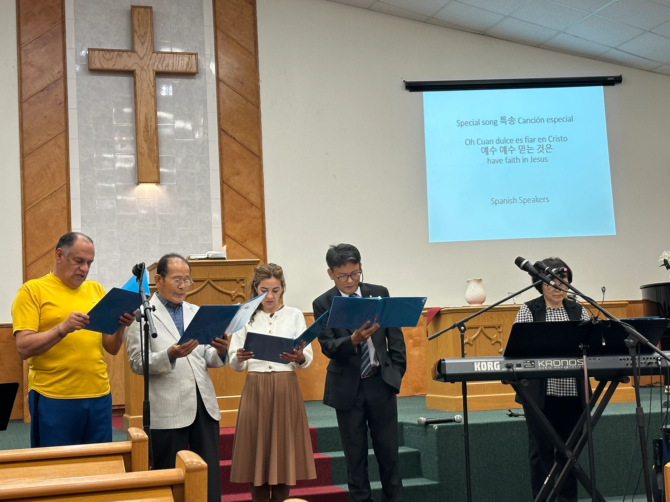 A diverse group of four singers and a keyboardist perform on stage in a church. A projection screen behind them displays song lyrics in three languages.