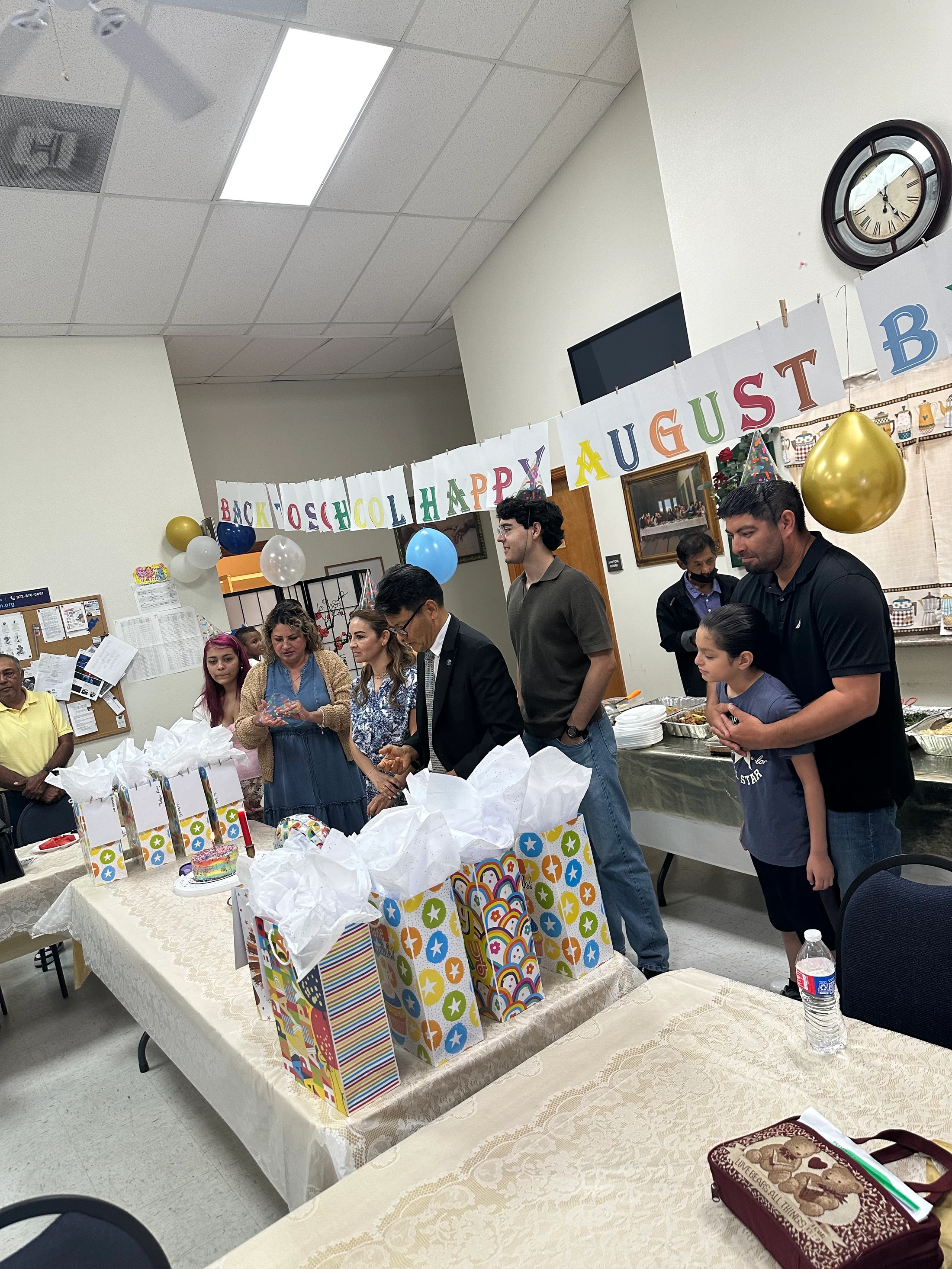 A group of adults and children gather in a room for a party. A table is lined with colorful gift bags and a small rainbow cake with a lit candle.