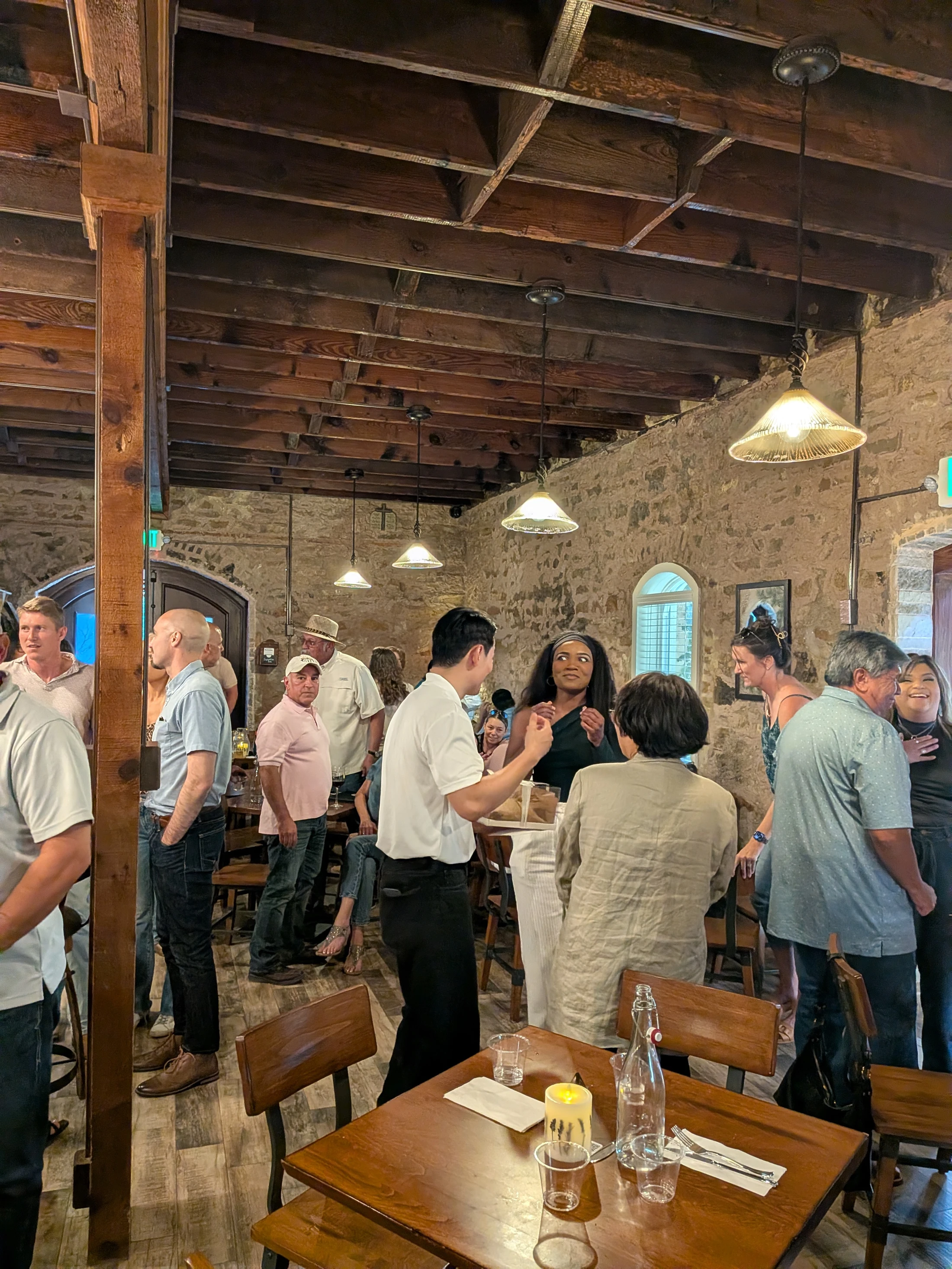 A diverse group of people socializes in a warmly lit restaurant with exposed stone walls and wooden ceiling beams, creating a lively, rustic atmosphere.