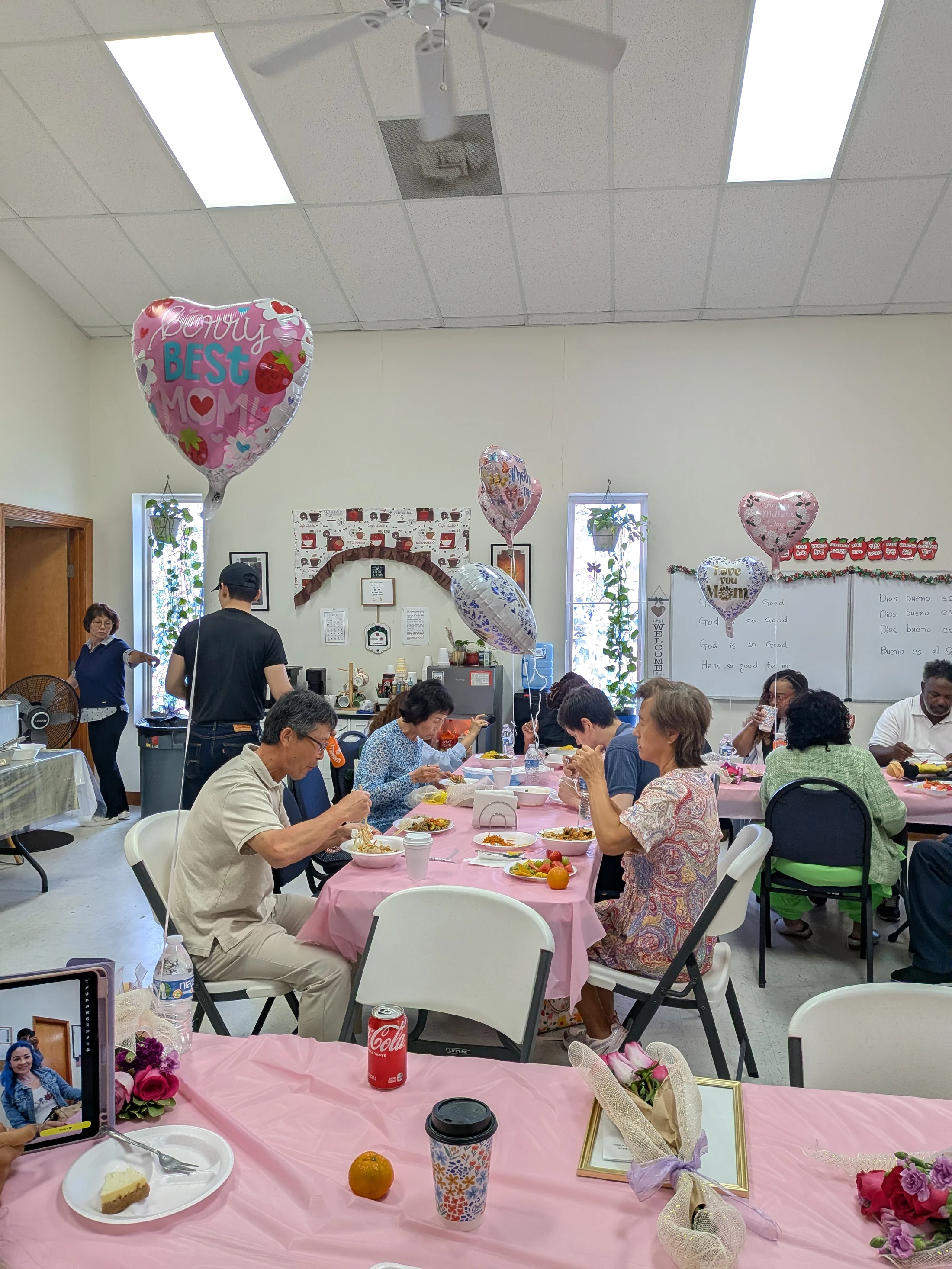 A diverse group of people sit at long tables with pink tablecloths, eating and celebrating Mother's Day in a community hall decorated with festive balloons.