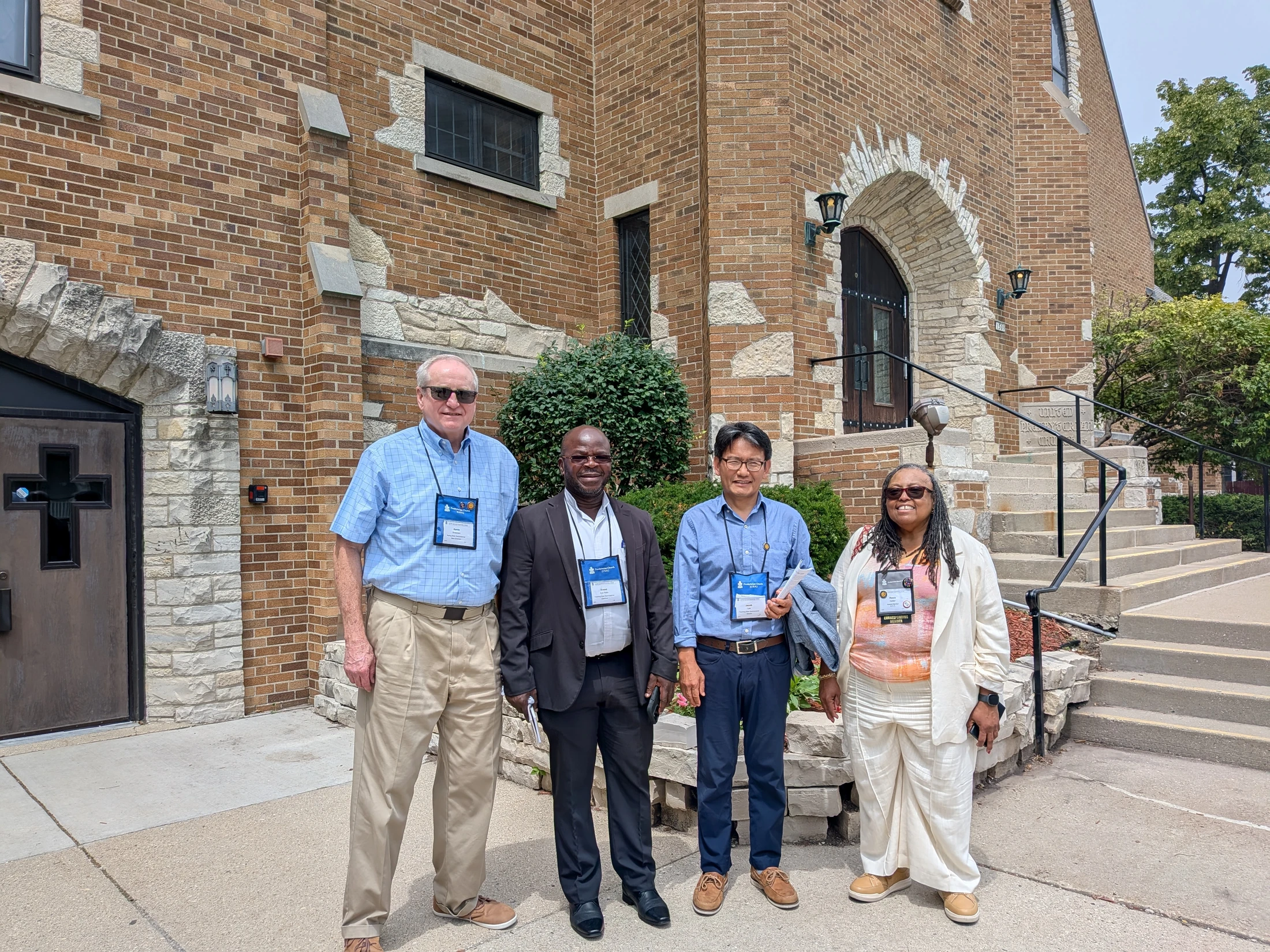 A diverse group of four people, three men and one woman, smile as they pose for a photo outside the entrance of a large brick and stone church building.