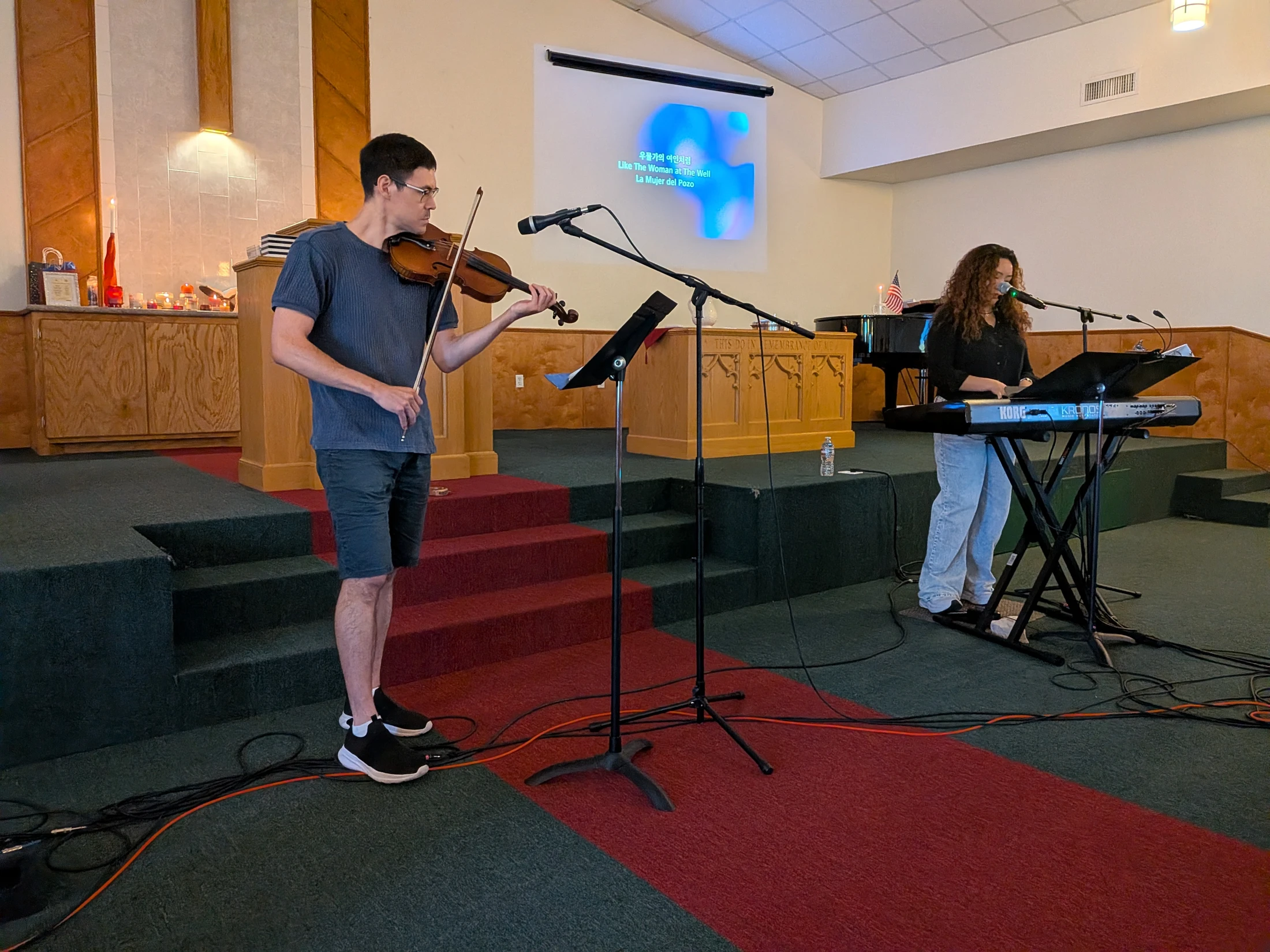 A man plays the violin and a woman plays an electronic keyboard on a stage in a church, with song lyrics projected on a screen behind them.