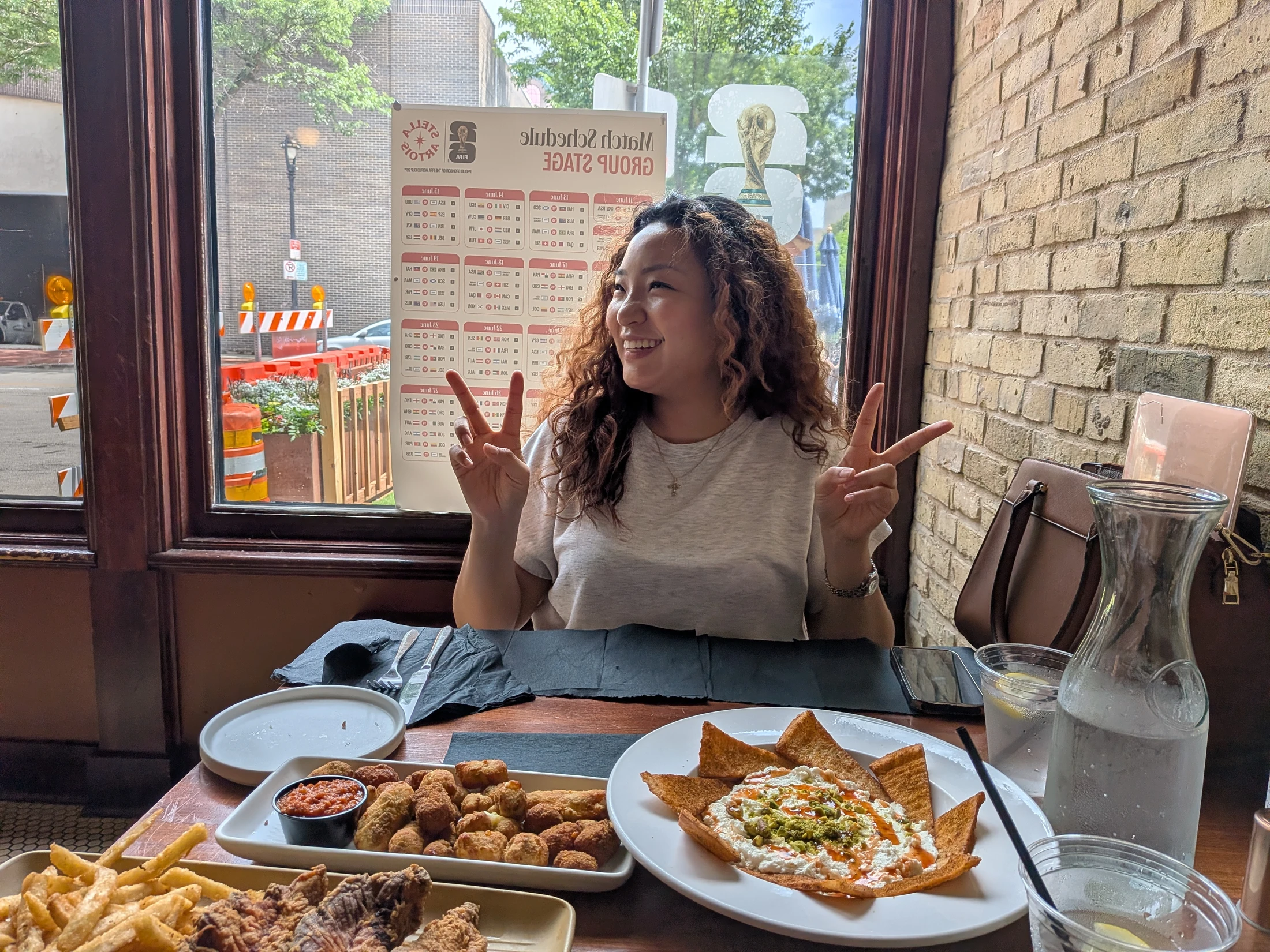 A smiling woman with curly hair makes peace signs with both hands while sitting at a restaurant table loaded with various appetizers next to a large window.