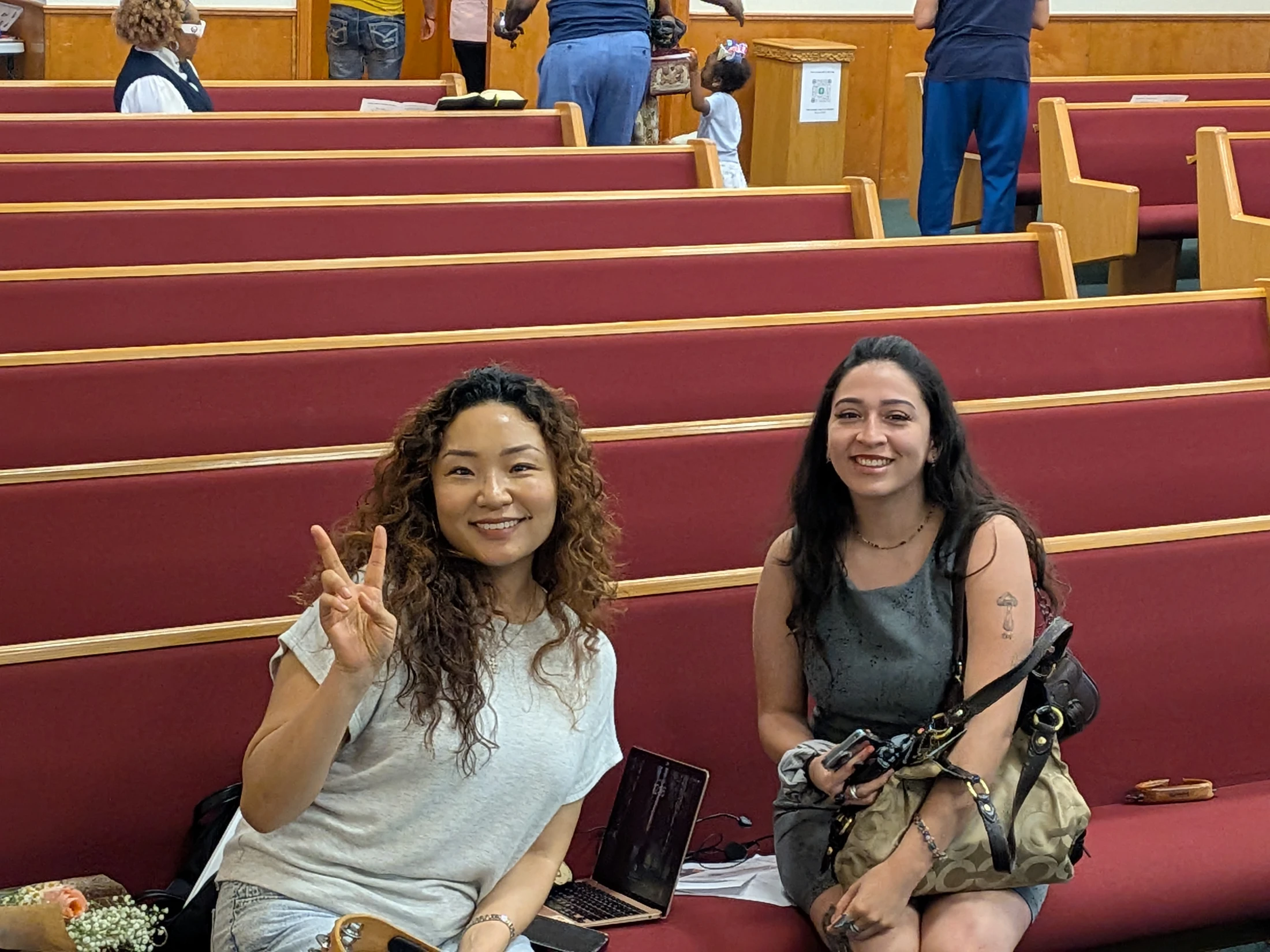 Two women smile for the camera while sitting on a red cushioned pew in what appears to be a church. One woman holds up a peace sign.