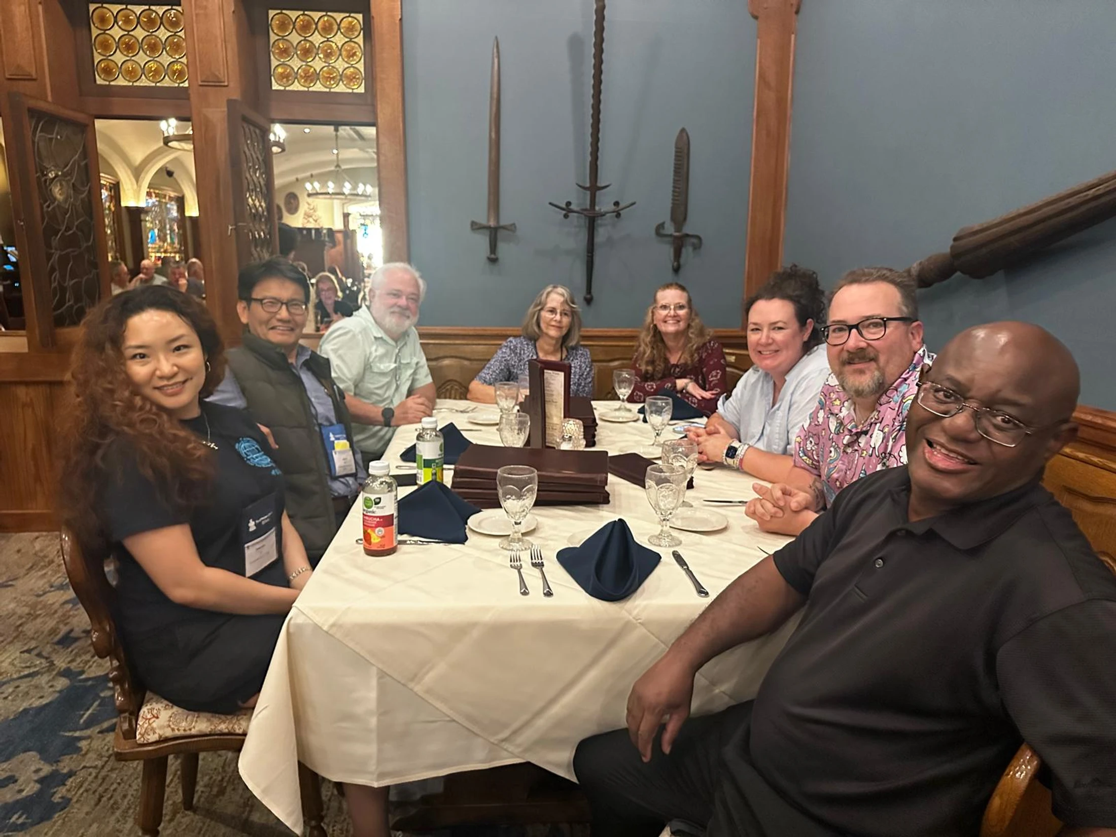 A diverse group of eight adults smile for the camera while seated at a long dining table in a restaurant with dark wood paneling and swords on the wall.