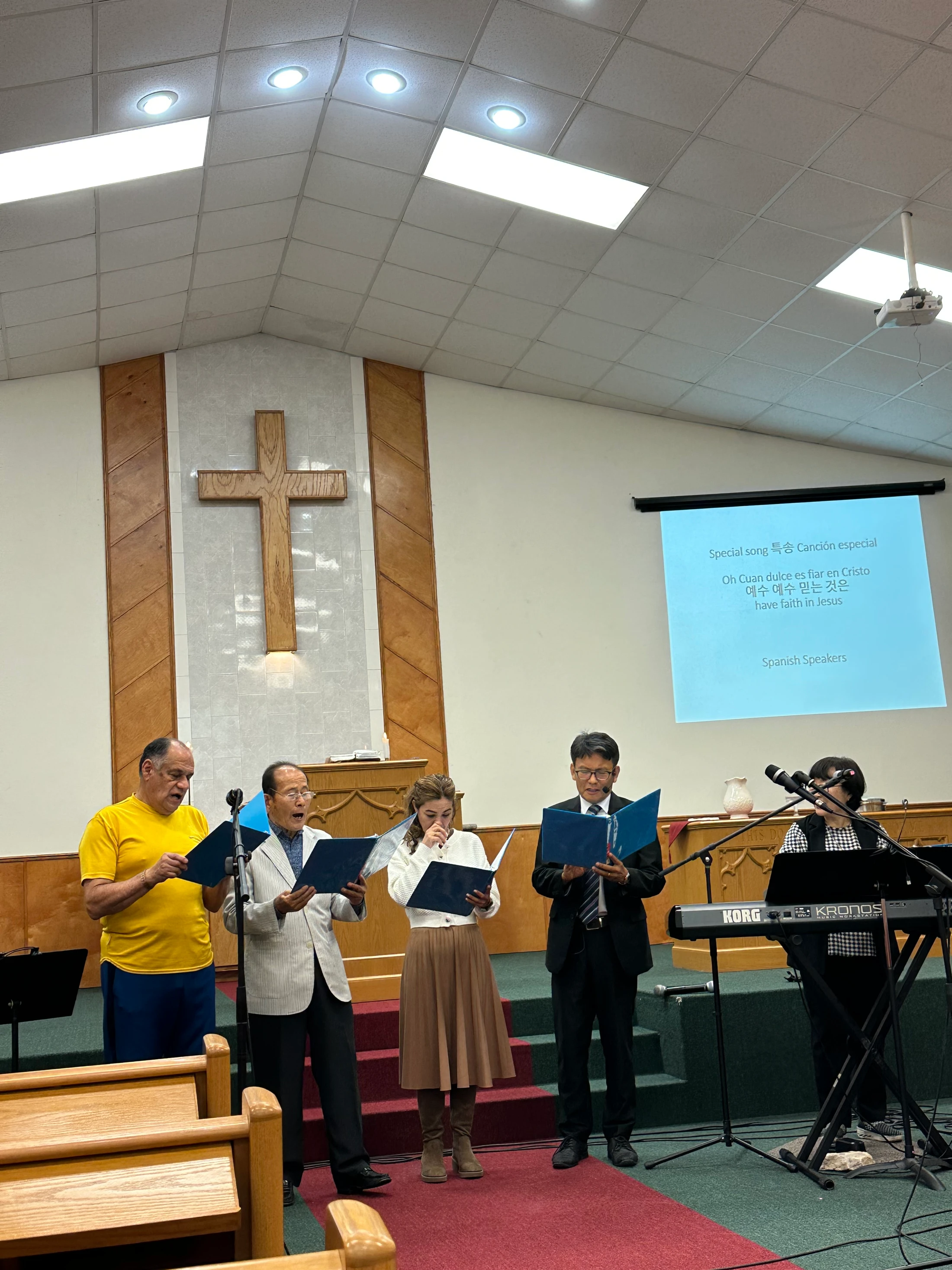 A diverse group of four singers and a keyboardist perform on a church stage in front of a cross. A screen displays song lyrics in multiple languages.
