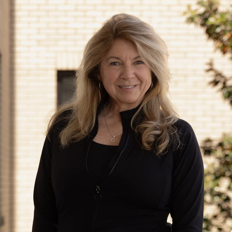 A woman with long, wavy blonde hair smiles warmly at the camera. She wears a black zip-up jacket and stands outdoors with a brick building in the background.