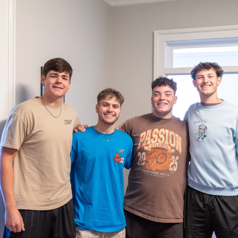 A group of four young men stand side-by-side with arms around each other, smiling for the camera in a brightly lit room.