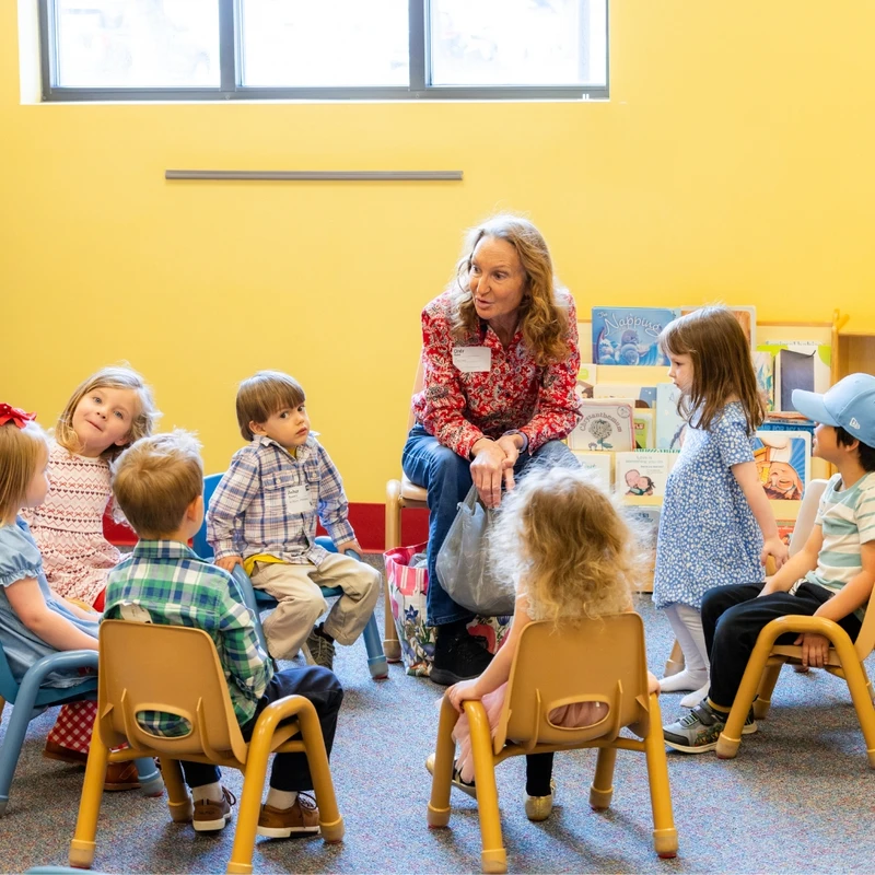 A woman sits in a circle with a group of young children in a classroom, talking to them as they listen attentively during what appears to be story time.