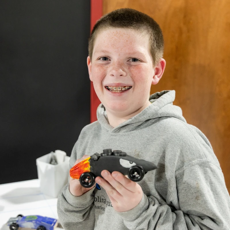 A smiling boy with freckles and braces wears a gray hoodie as he proudly holds up a small, black, handmade wooden race car painted with flames.