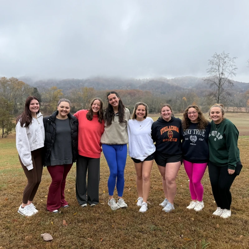 A group of eight young women stand side-by-side, smiling for a photo in a field with foggy mountains and autumn trees in the background.