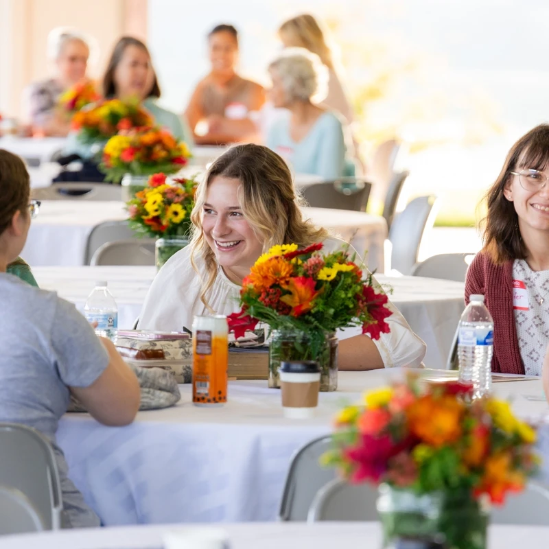 A group of women smile and talk while seated at tables with white cloths and colorful autumn flower bouquets during an outdoor social event.