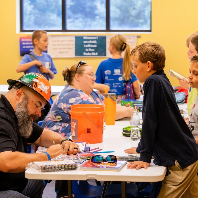 A bearded man in a decorated orange hard hat sits at a table and signs something for a young boy who is watching him intently in a classroom setting.