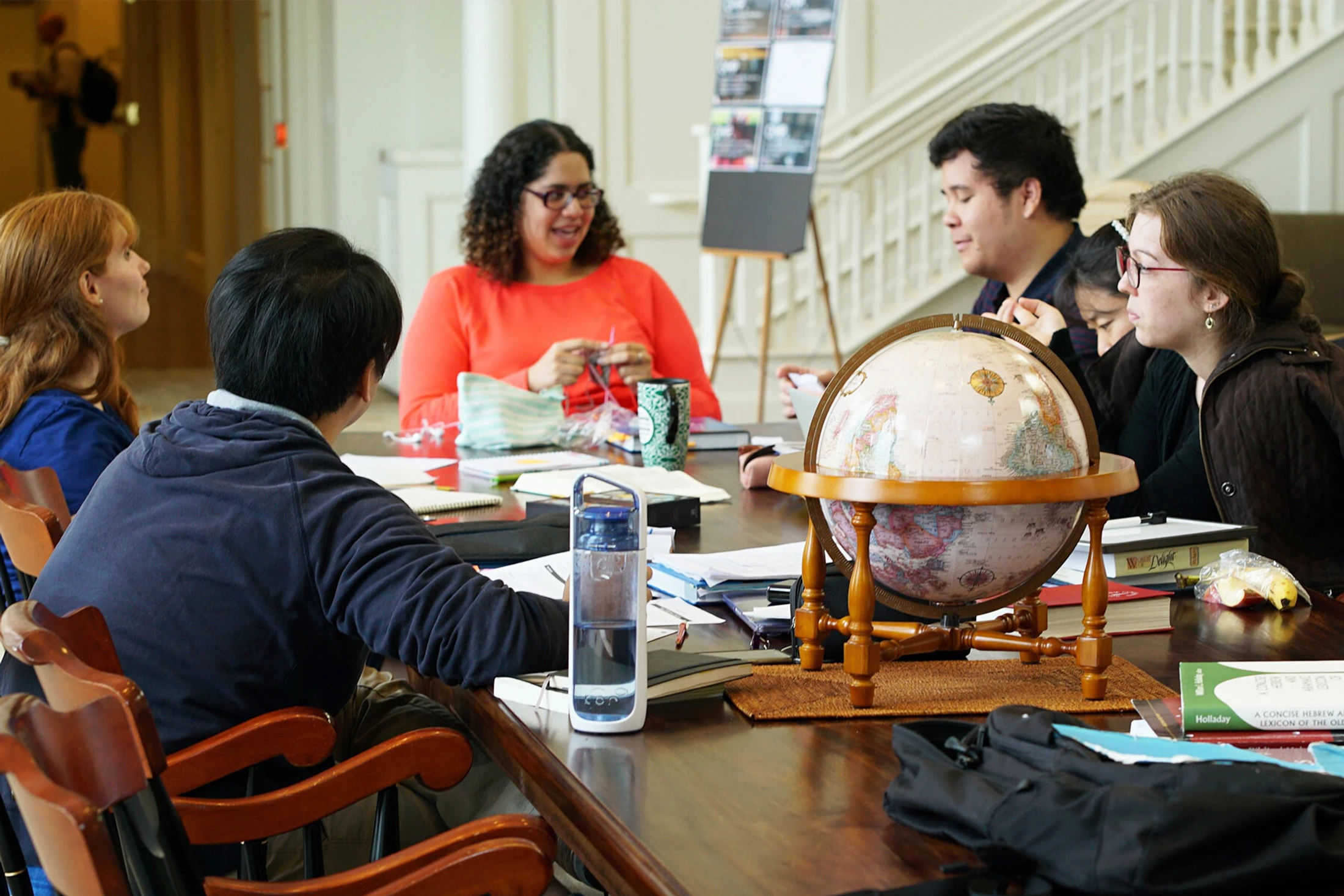 A diverse group of five people sits around a long wooden table, engaged in a discussion. On the table are books, papers, and a decorative globe.