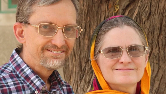 A middle-aged man in a plaid shirt and a woman wearing a colorful headscarf smile for a portrait in front of a large tree.