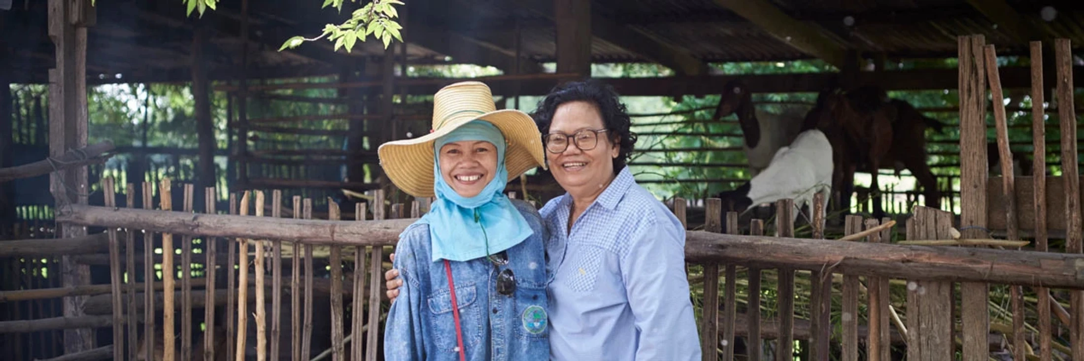 Two smiling women stand together in front of a rustic wooden goat pen. One wears a straw hat and the other wears glasses. Goats are visible behind them.