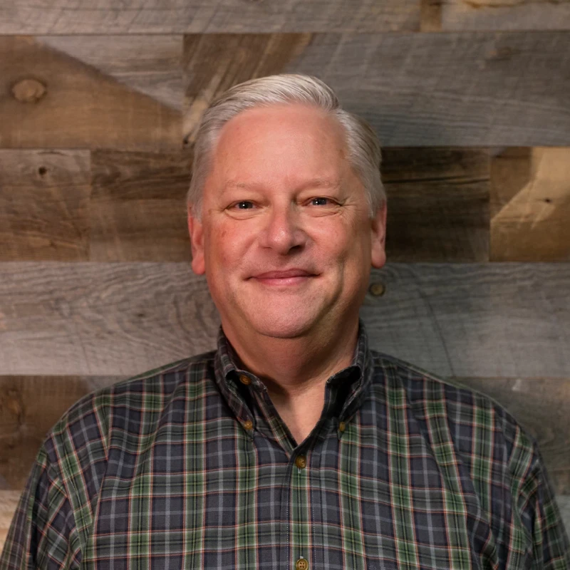 A smiling middle-aged man with grey hair, wearing a green and grey plaid shirt, poses for a headshot against a rustic wood-paneled background.