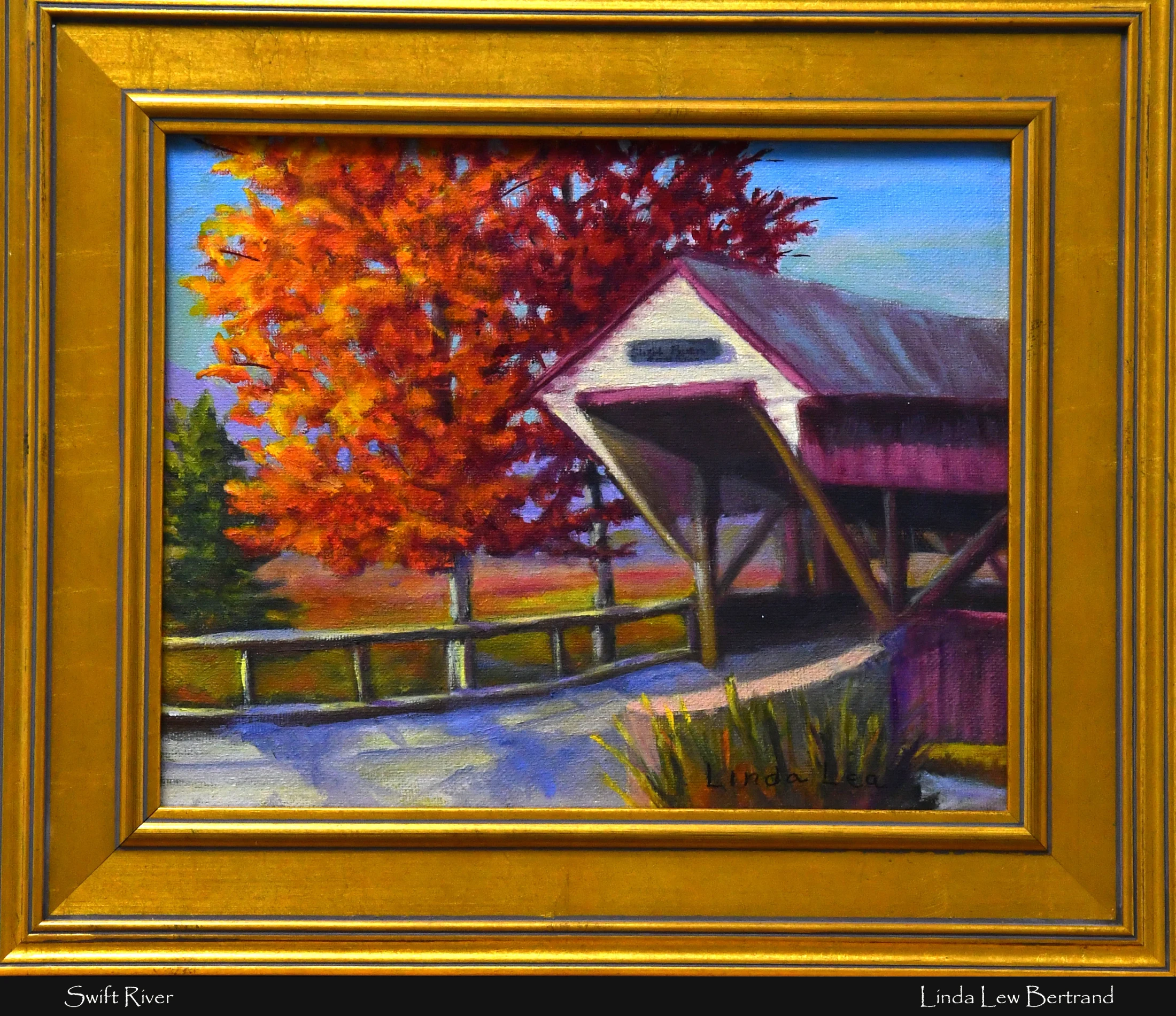 An oil painting of a covered bridge next to a tree with vibrant orange autumn leaves. The artwork, titled 'Swift River', is set in an ornate gold frame.