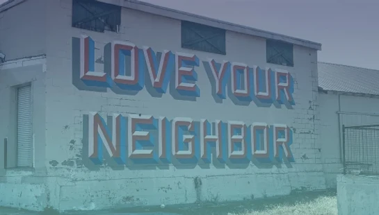A mural on the side of a weathered building reads 'Love Your Neighbor' in large, 3D-style block letters in red, white, and blue.