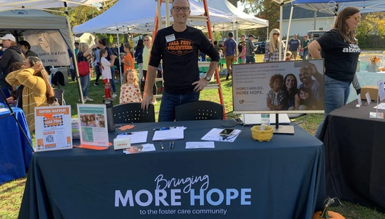 A male volunteer stands smiling behind a Fostering Hope booth at an outdoor fall festival, ready to share information about the foster care community.