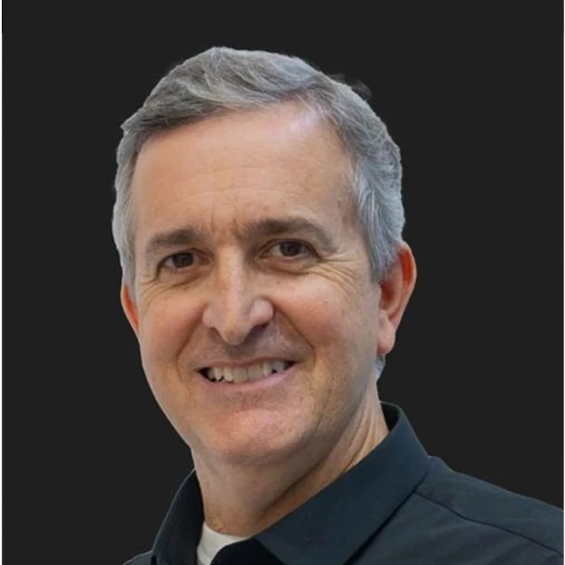 A close-up headshot of a middle-aged man with short, graying hair. He is smiling at the camera, wearing a black collared shirt over a white t-shirt.