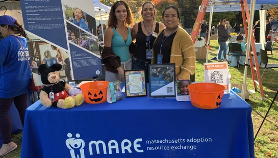 Three women smile behind a booth for the Massachusetts Adoption Resource Exchange (MARE) at a sunny outdoor event with fall foliage in the background.