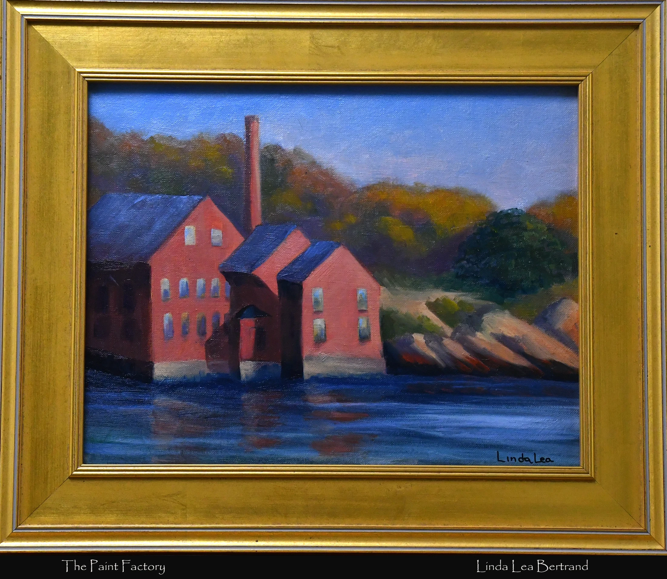 A framed oil painting called 'The Paint Factory' depicting red industrial buildings on the edge of a body of water, with a forested hill in the background.