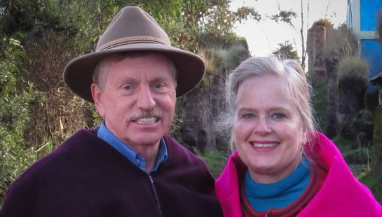 A smiling man and woman stand side-by-side outdoors. The man wears a brown hat and a purple poncho, and the woman is wrapped in a bright pink poncho.