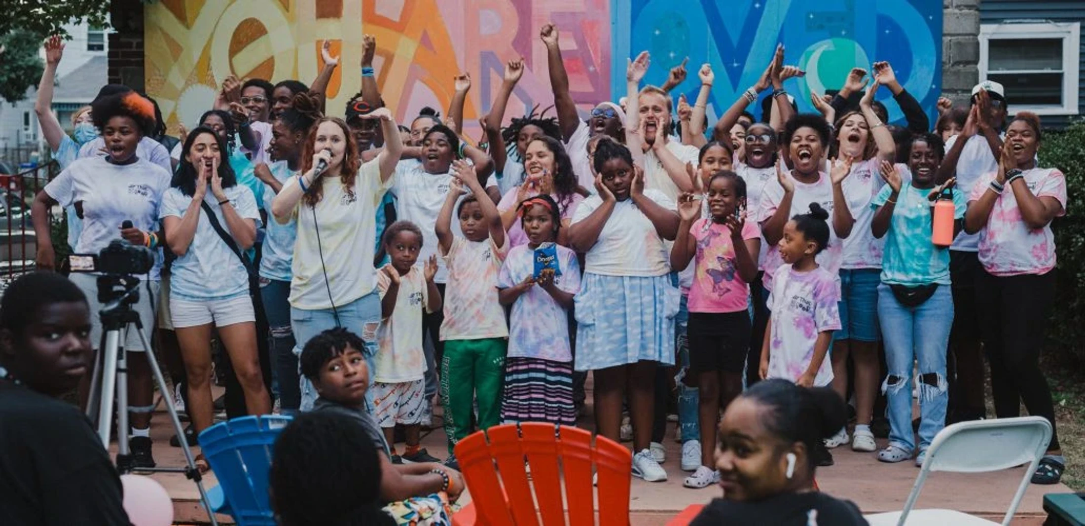A large, diverse group of adults and children cheer with their hands in the air while standing in front of a colorful mural during an outdoor event.