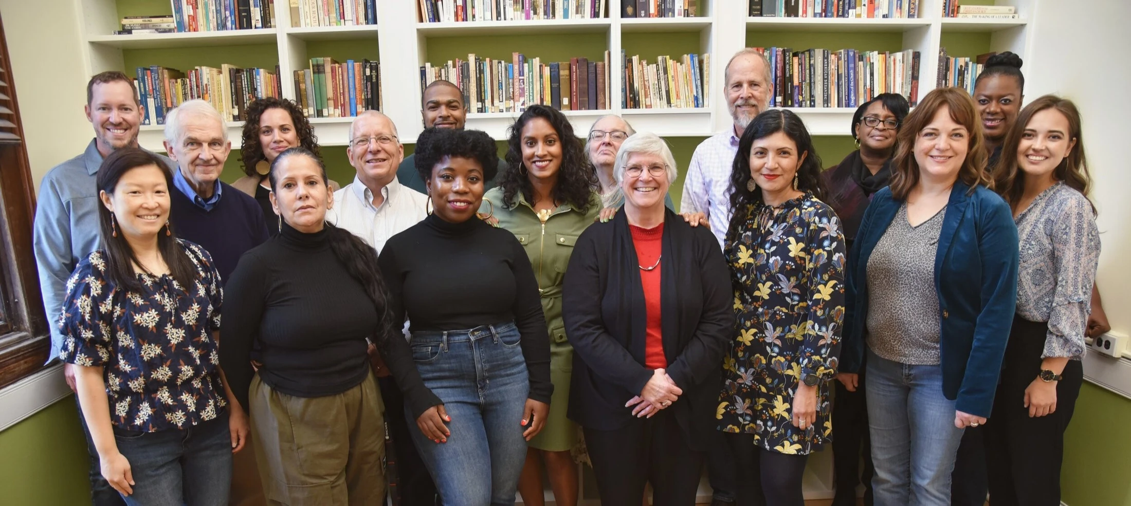 A diverse group of about fifteen people, men and women of various ages, stand together smiling for a photo in front of green and white bookshelves.