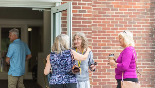 Three senior women stand outside a brick building entrance, smiling and talking. Two embrace while another holds a coffee cup. A man enters the building.