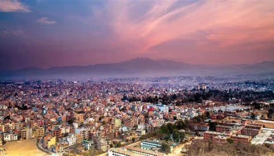 A sprawling, dense cityscape of Kathmandu, Nepal, viewed from above at sunrise, with mountains in the misty background under a pink and purple sky.