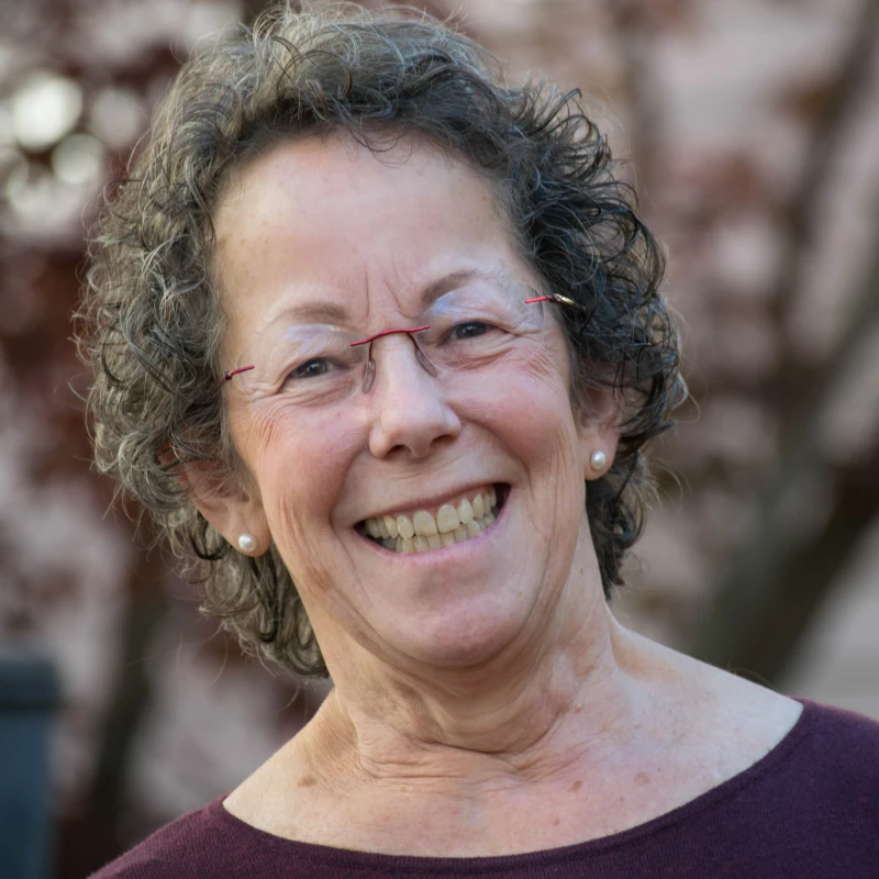 A close-up portrait of an older woman with short, curly gray hair. She is smiling widely, wearing rimless glasses and pearl earrings.