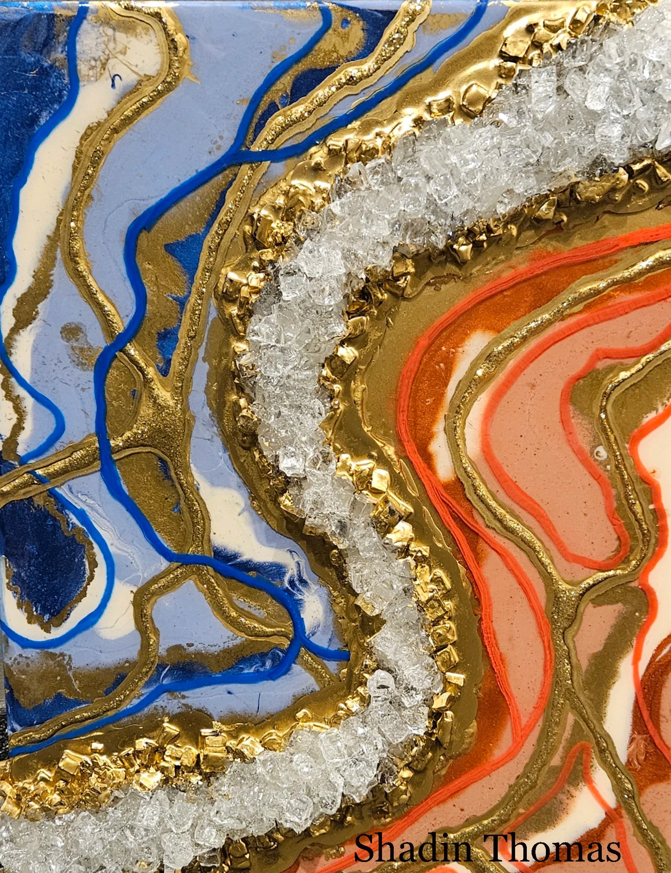 A close-up of a mixed-media abstract painting with blue and orange swirls, outlined in gold and separated by a textured band of gold leaf and clear crystals.