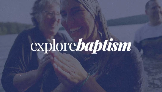 A smiling young woman with wet hair has her hands clasped during an outdoor water baptism, with text that reads 'explorebaptism'.