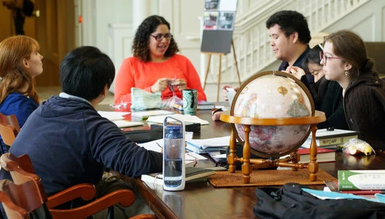A diverse group of five people sits around a long wooden table, engaged in a discussion. On the table are books, papers, and a decorative globe.