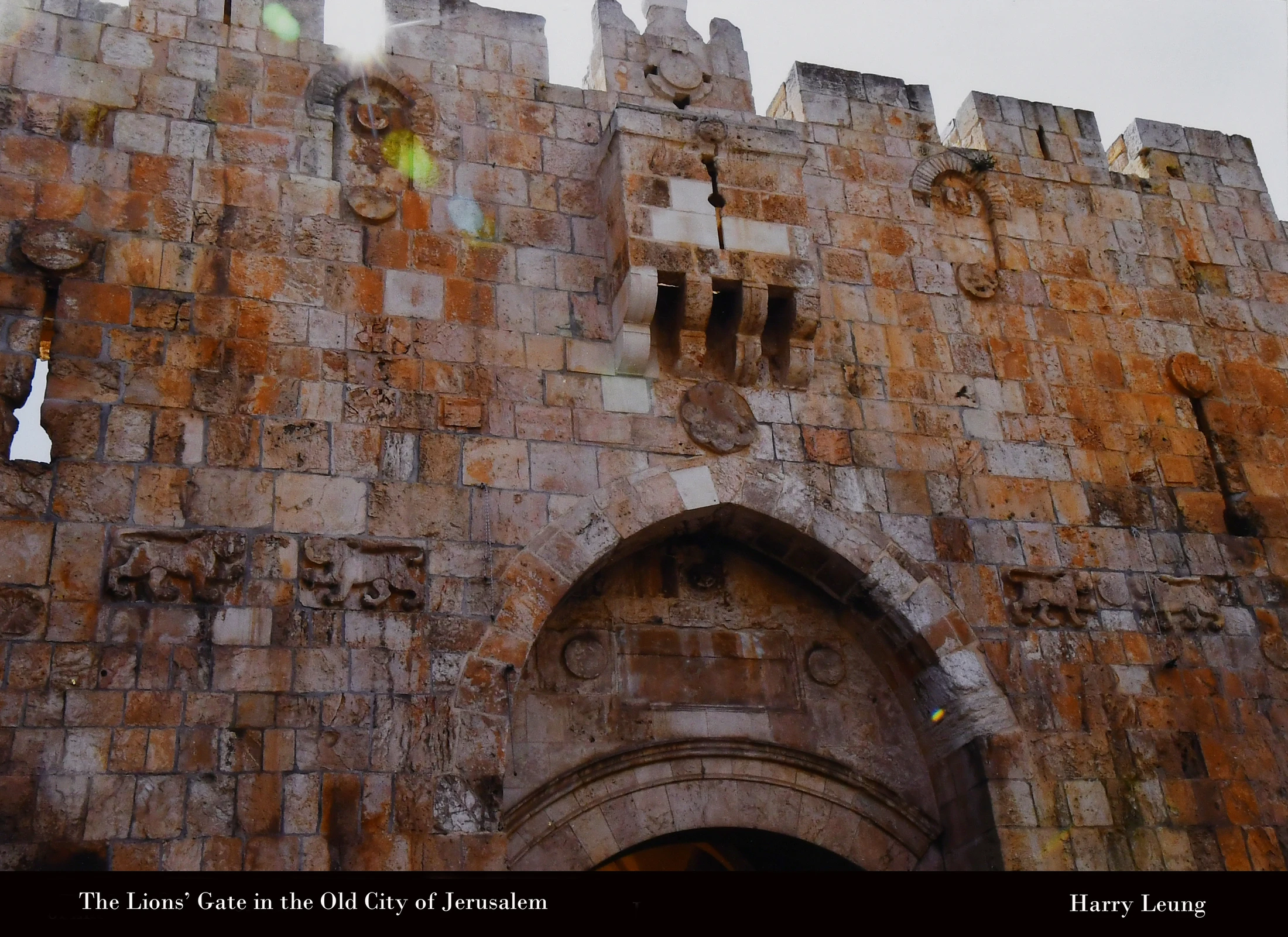 A close-up of the weathered, tan stone wall of the Lions' Gate in Jerusalem, showing the main arch, crenellations, and carvings with a sun flare at the top.