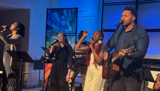 Four singers perform on a blue-lit stage. A man on the right plays an acoustic guitar while he and three women sing passionately into microphones.