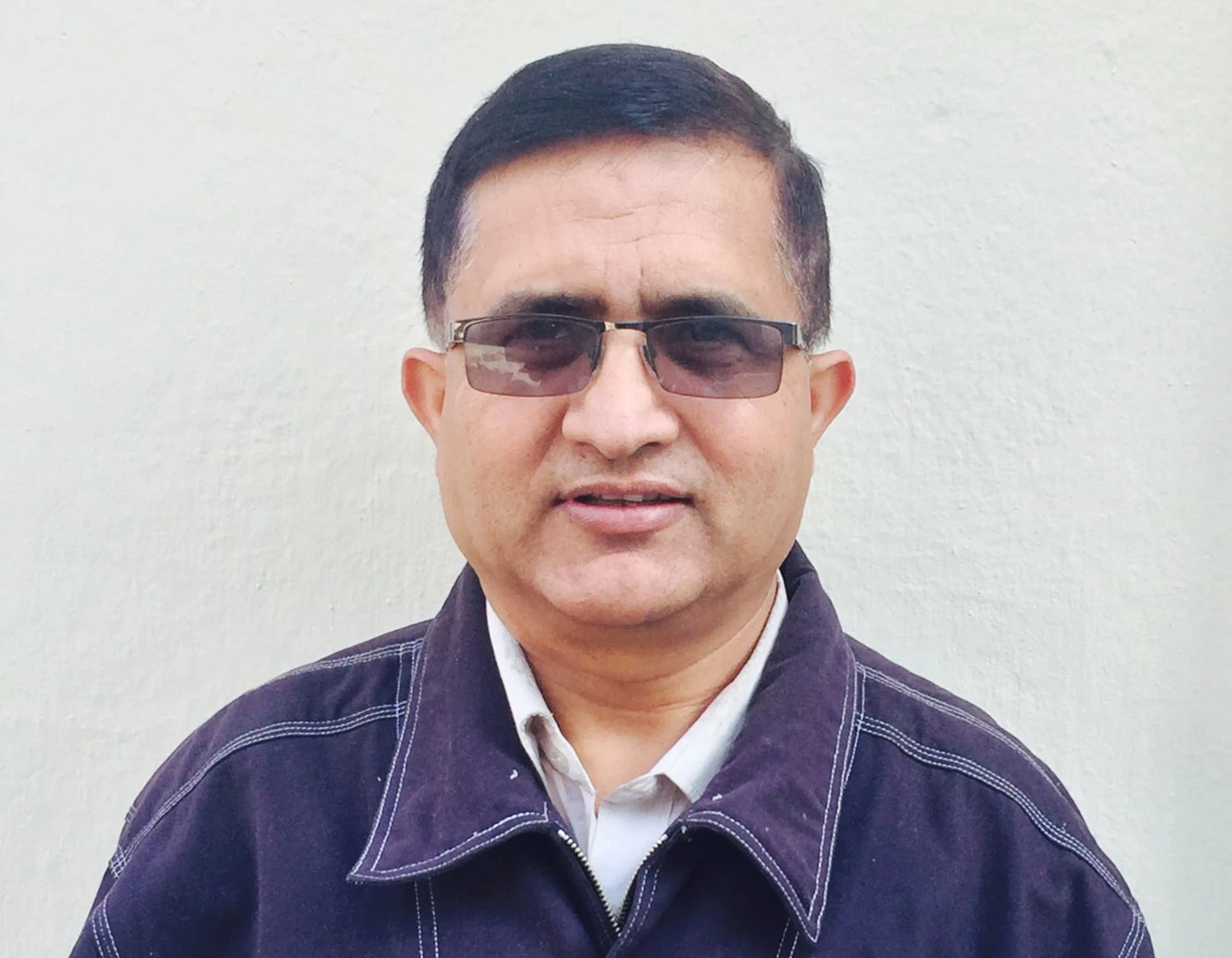 A head-and-shoulders portrait of a middle-aged man with short dark hair. He wears tinted glasses, a collared shirt, and a dark jacket against a plain wall.