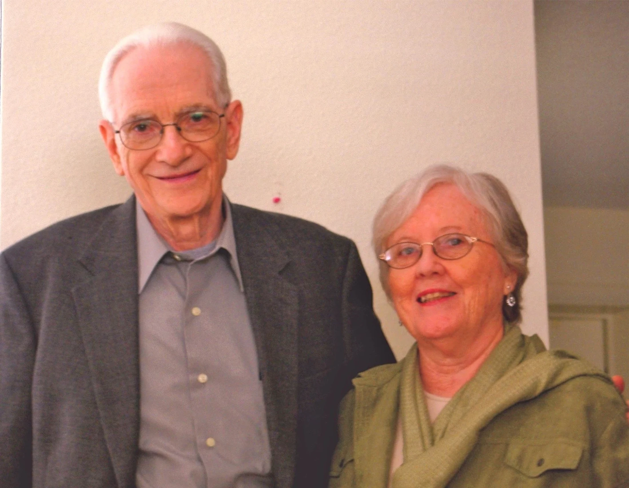 A waist-up portrait of an elderly man and woman standing side-by-side. He wears a grey blazer and she wears a green jacket. Both have white hair and glasses.