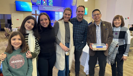A group of seven people of various ages smiling for a photo inside a church hall during a gathering, with stained glass windows visible in the background.
