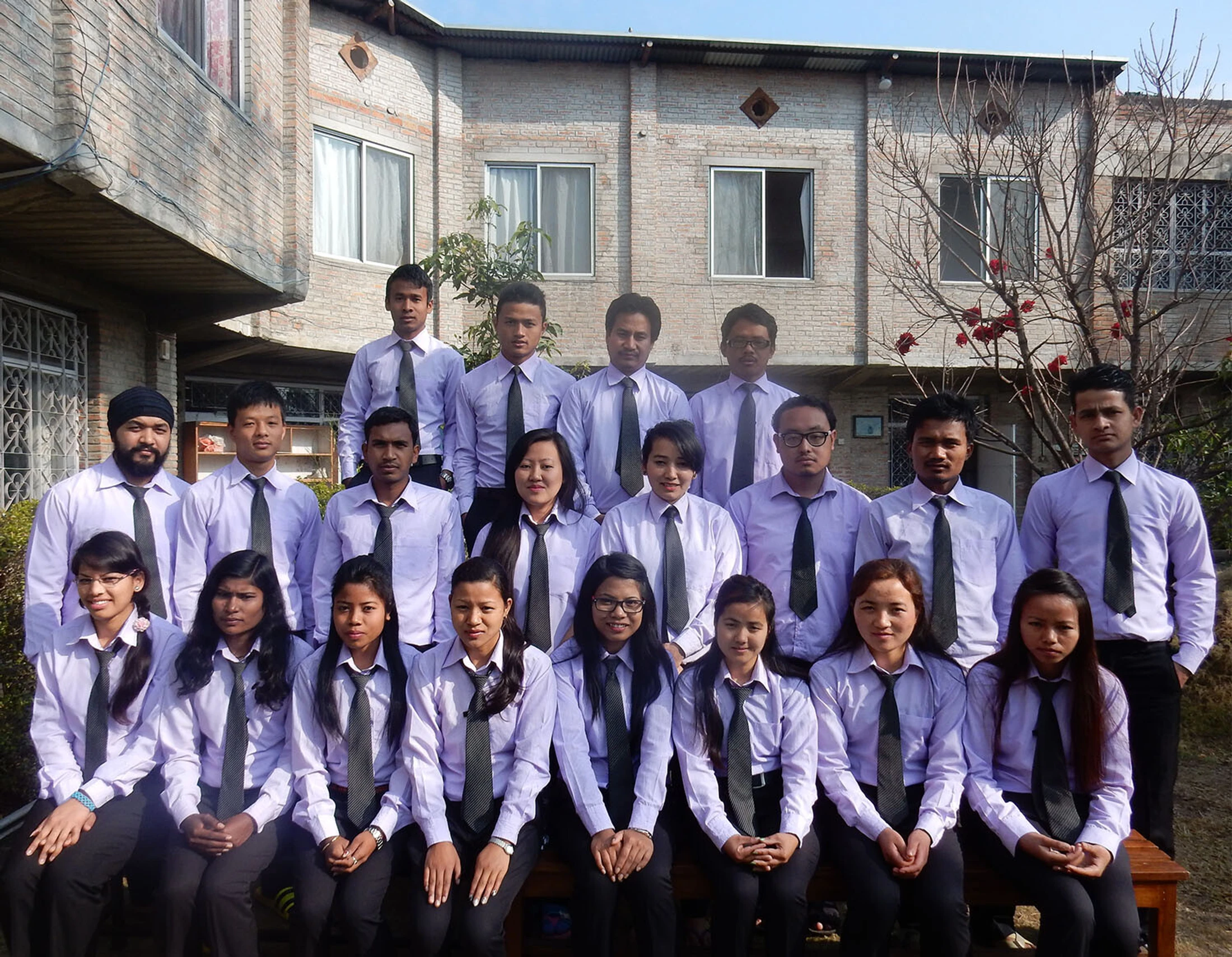 A group of male and female students in matching purple shirts and dark ties pose for a formal portrait outdoors in front of a brick building.