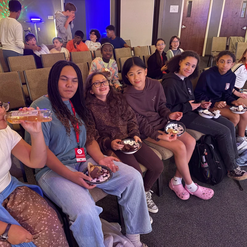 A diverse group of middle and high school students sit in rows of chairs, smiling and eating cake from paper plates during a social gathering.