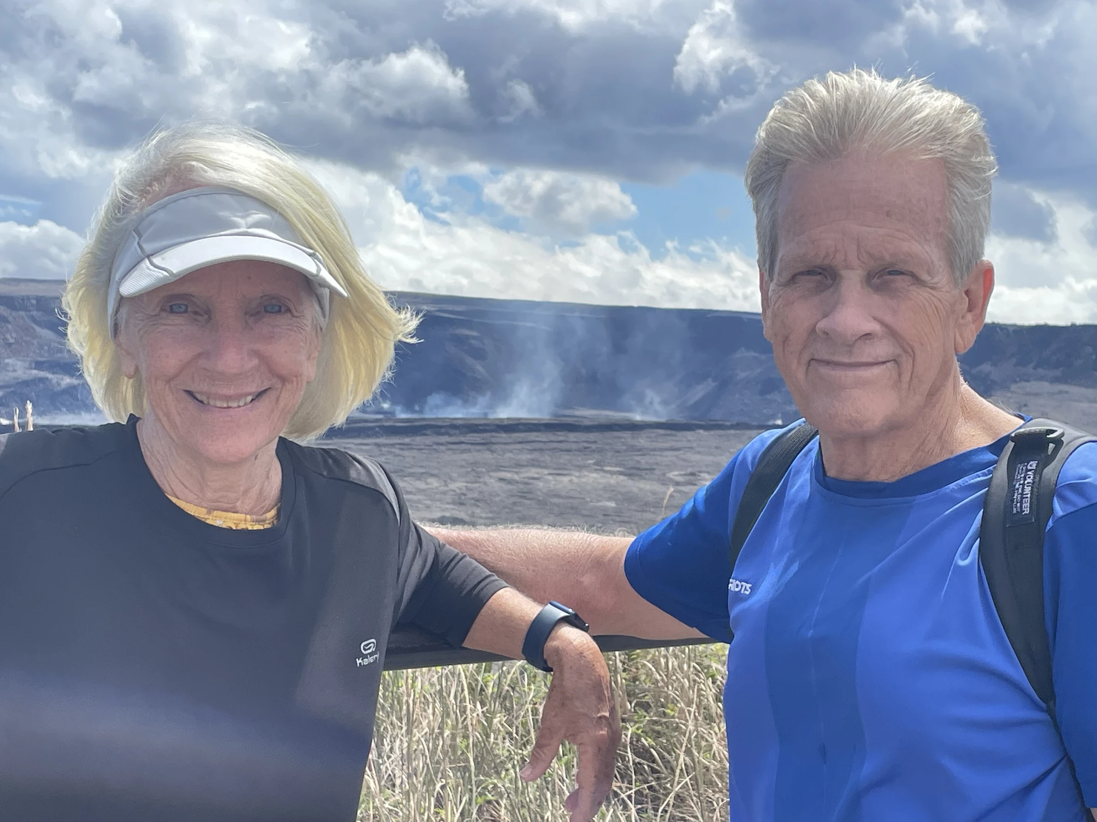 An older man and woman in athletic wear smile for the camera, posing in front of a vast volcanic crater with steam rising from it under a cloudy sky.