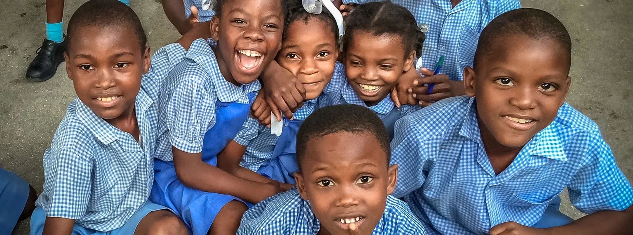 A group of young Black schoolchildren in blue and white checkered uniforms smile and laugh together while posing for a photo.