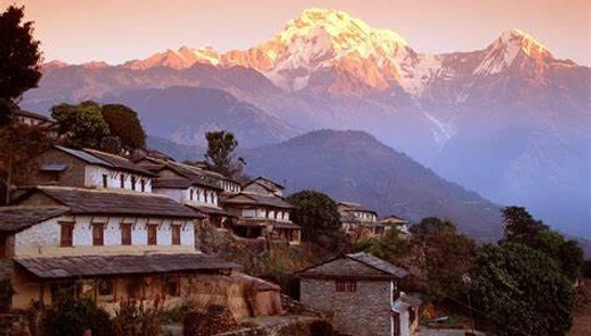 A tranquil village on a terraced hillside with traditional houses, overlooking massive, snow-capped mountains glowing golden at sunset.