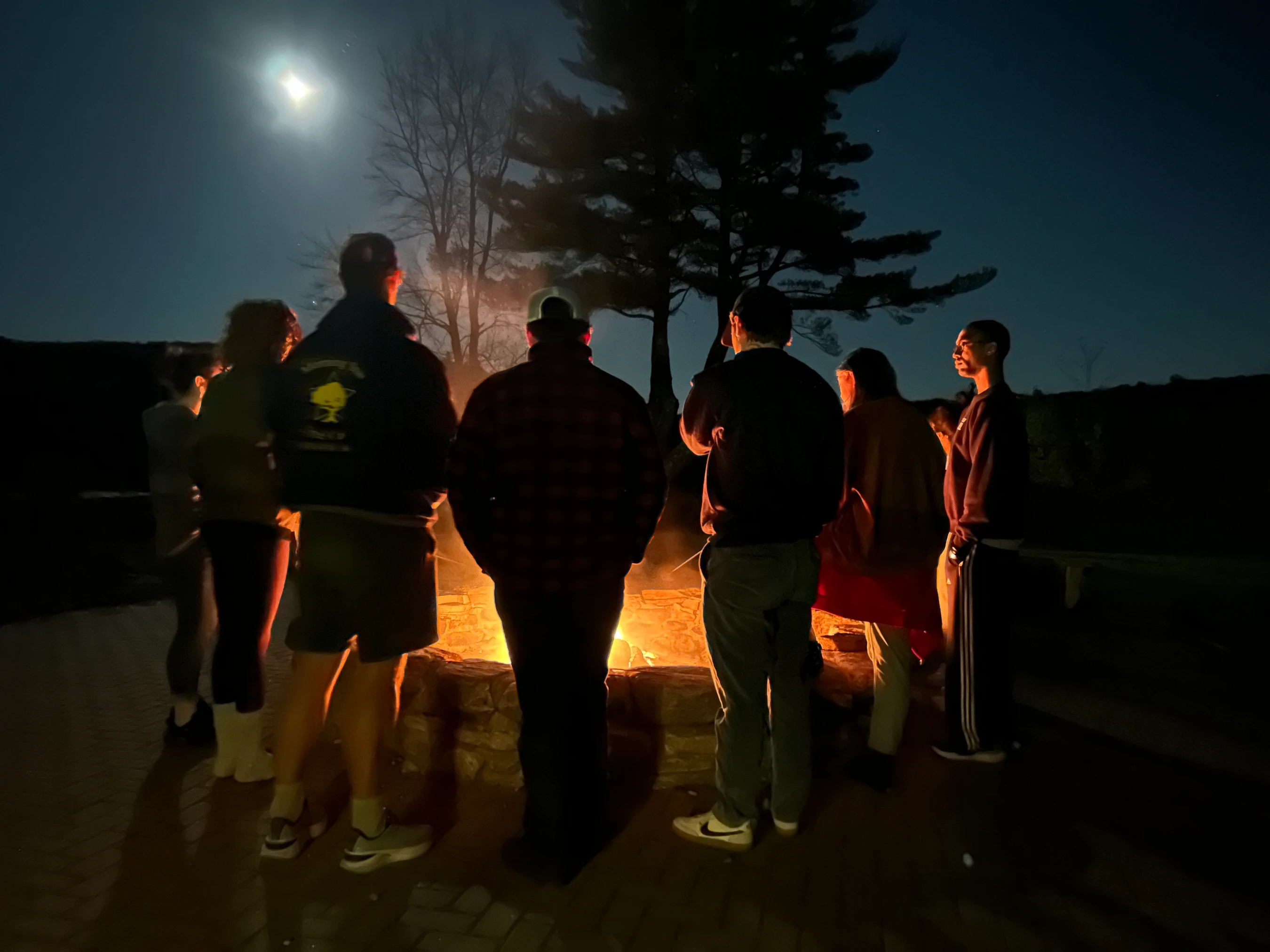 A group of people stand in a circle around a glowing fire pit on a brick patio at night. They are silhouetted against the fire under a bright moon.
