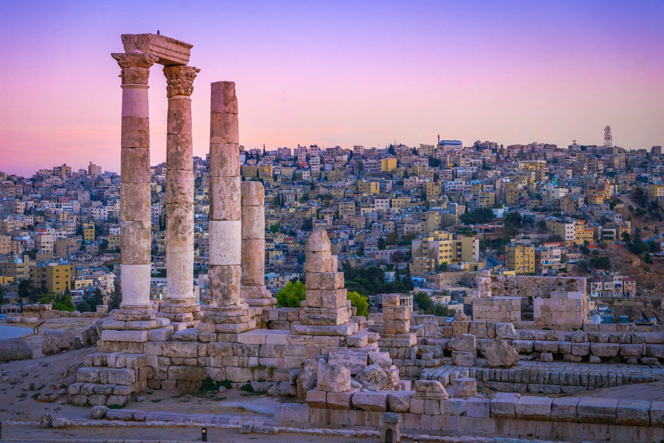 The ancient ruins of the Temple of Hercules at the Amman Citadel, with its tall stone columns, overlook the sprawling city of Amman under a vibrant sunset.