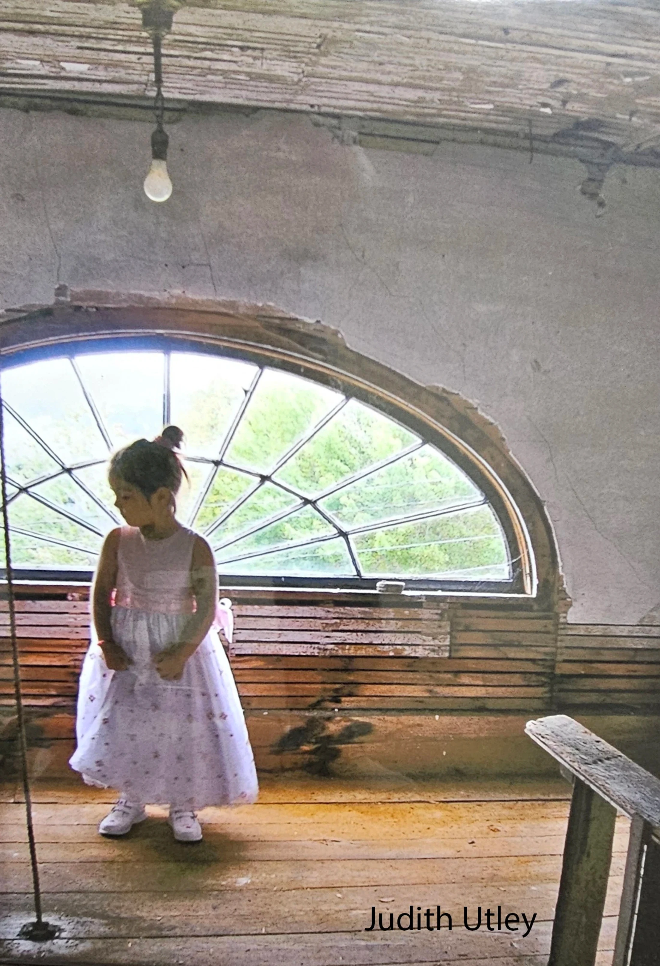 A young girl in a fancy light purple dress stands in a rustic, unfinished room with a large, arched window behind her and a bare lightbulb overhead.