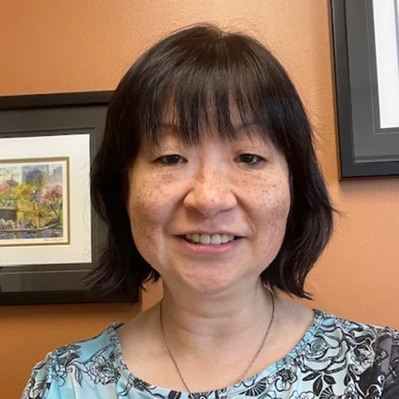 A headshot of a smiling East Asian woman with shoulder-length black hair, bangs, and freckles, wearing a patterned top in front of an orange wall.